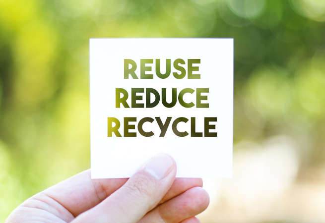 Eine Hand die einen weißen Zettel mit der Aufschrift „Reuse, Reduce, Recycle“ vor einen grünen Hintergrund hält